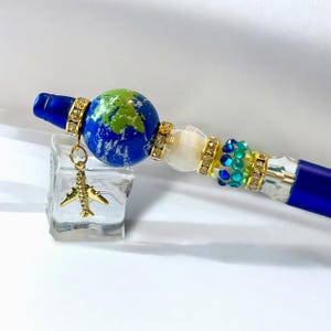 May include: A blue pen with a globe design and a gold airplane charm. The pen features a blue barrel, a globe bead with green and blue details, and decorative gold and crystal accents. The pen is approximately 6 inches long.
