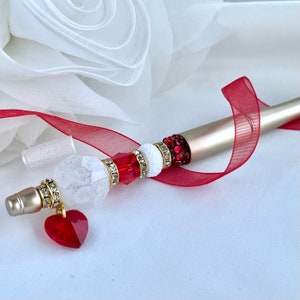 May include: A silver pen with a white and red beaded design. The pen has a heart-shaped charm at the end. The pen is lying on a white surface with a red ribbon.