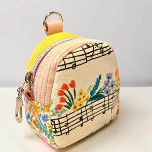 May include: A small, structured pouch featuring a cream-colored canvas front with black musical staff lines and embroidered colorful flowers. The sides are made of yellow and light pink corduroy fabric, and it includes a silver metal zipper and a top ring.