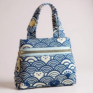 May include: A small, blue and white patterned handbag with a wave and cat design. The bag has a light blue zipper and a gold-colored zipper pull. The handles are attached with a gold-colored button.