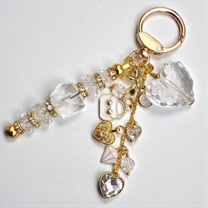 Op de afbeelding: Een goudkleurige sleutelhanger met een heldere, facetgeslepen kristallen toverstaf en diverse bedels. Bedels zijn onder meer een hart, een diamant en een strik. De sleutelhanger heeft een gouden sluiting en is versierd met heldere kristallen.