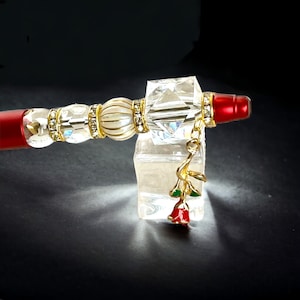 May include: A gold pen with a red tip, clear crystal accents, and a white pearl bead. A gold charm with a red rose and green leaves hangs from the pen.