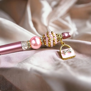 K&ouml;nnte beinhalten: Ein rosa-goldener Perlenstift mit einem Anh&auml;nger in Form einer kleinen Handtasche. Der Handtaschenanh&auml;nger hat eine rosa Schleife und Strasssteine.