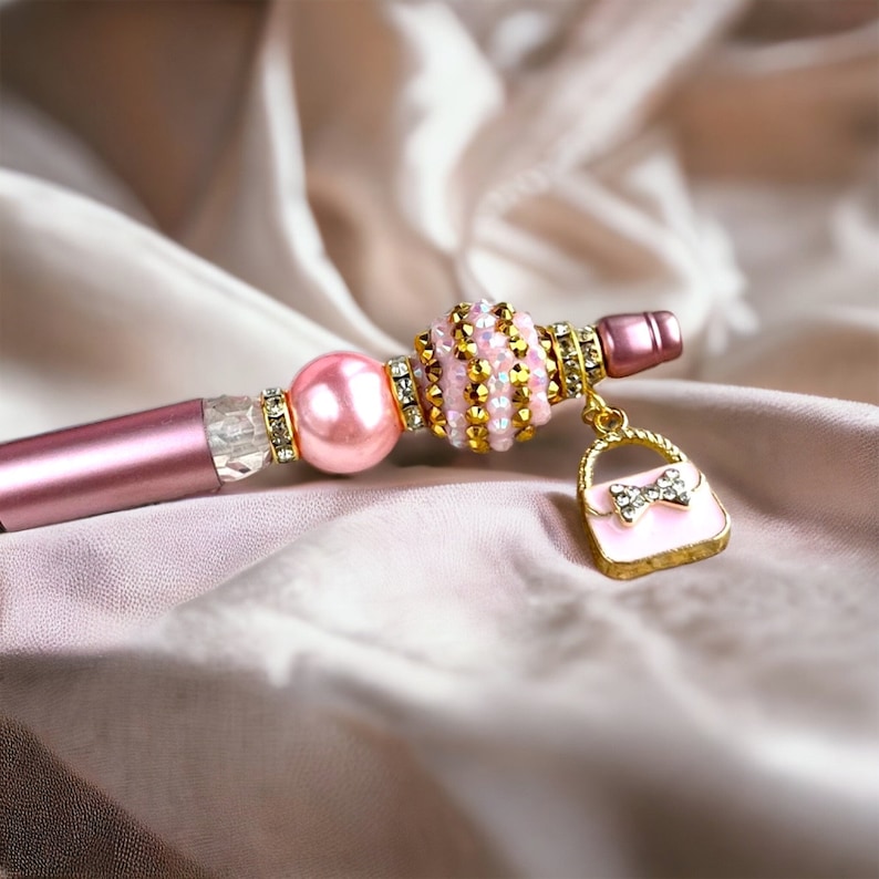 K&ouml;nnte beinhalten: Ein rosa-goldener Perlenstift mit einem Anh&auml;nger in Form einer kleinen Handtasche. Der Handtaschenanh&auml;nger hat eine rosa Schleife und Strasssteine.
