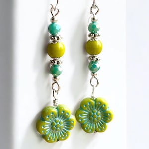 May include: A pair of dangle earrings featuring a floral design. The earrings have a silver hook, with a strand of green and yellow beads. The bottom of the earrings have a green flower with a turquoise center.