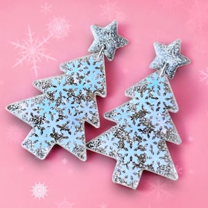 May include: A pair of silver glitter Christmas tree earrings with a blue snowflake design. The earrings are made of resin and have a star charm at the top.