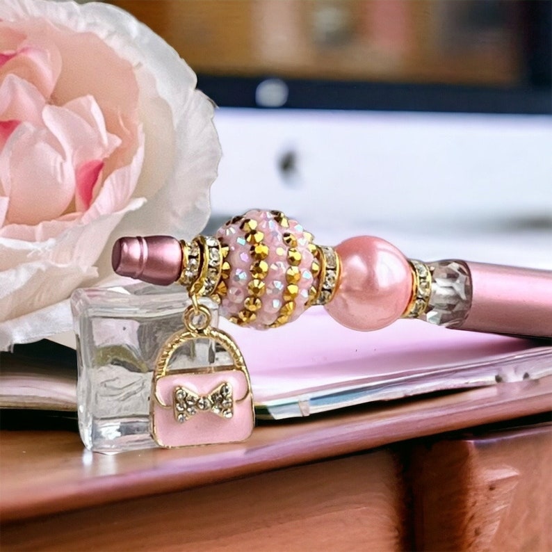 K&ouml;nnte beinhalten: Ein rosa und goldener Stift mit einer Perlen- und Strassverzierung. Der Stift hat einen Anh&auml;nger in Form einer rosa Handtasche mit einer Strassschleife.