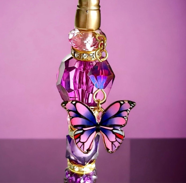 May include: A purple glass bottle stopper with a gold top and a pink and blue butterfly charm. The bottle stopper is decorated with rhinestones and crystals.