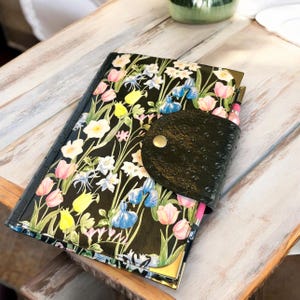 May include: A black leather journal with a floral pattern and a flap closure. The journal has a gold accent on the edge and a gold button on the flap.