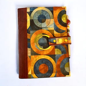 May include: A brown leather-bound journal with a geometric pattern of circles and squares in shades of orange, yellow, blue, and gray. The journal has a gold metal ring and a brown leather strap with gold accents.