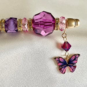 May include: A purple and pink crystal pen with a gold butterfly charm. The pen has a clear crystal barrel with purple and pink beads and a gold cap.
