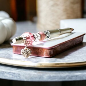 May include: A silver pen with pink and clear crystal beads and a heart-shaped charm. The pen is resting on a white and pink rectangular box.