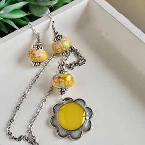 May include: A yellow and white floral necklace and earrings set with a silver chain and a yellow flower pendant.