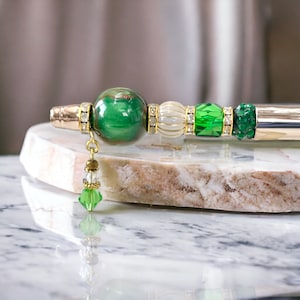 Può includere: Una penna verde e oro con un grande ciondolo di pietra preziosa verde, perline bianche e dorate e dettagli in cristallo verde. La penna ha un corpo in metallo argentato e un piccolo ciondolo di cristallo verde.