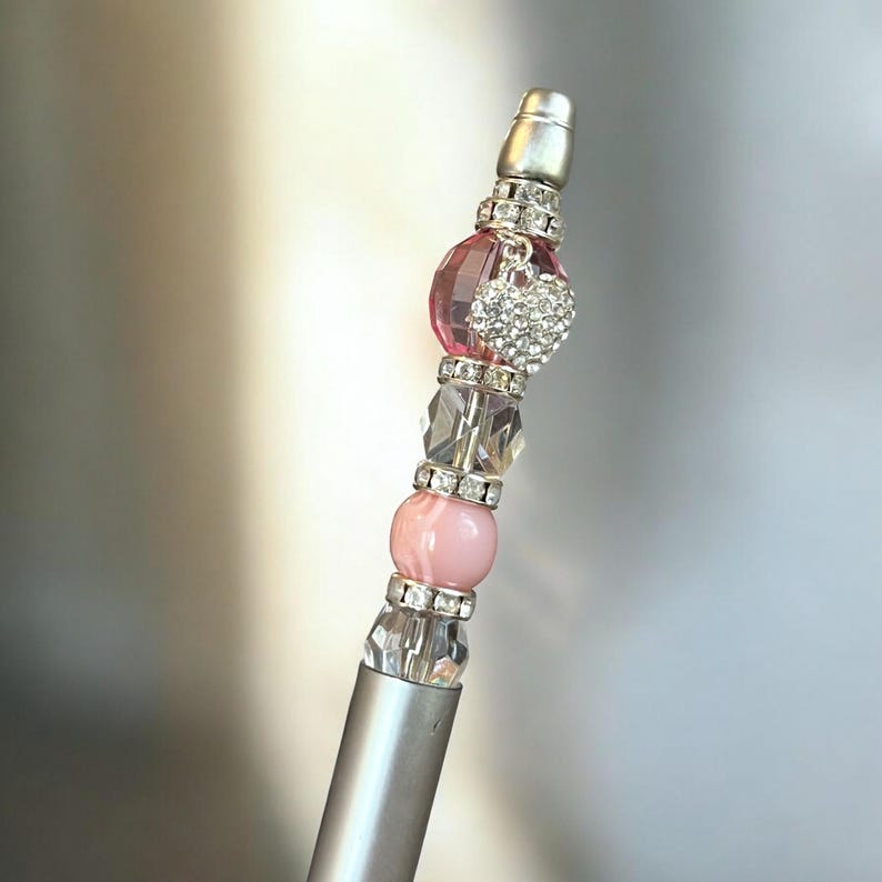 May include: A silver pen with a pink and clear crystal bead charm. The charm has a small heart-shaped pendant with rhinestones.