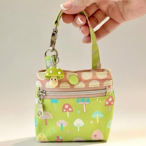 May include: A small, square coin purse with a green mushroom pattern and a zipper closure. The purse has a small handle and a mushroom-shaped charm attached. The top of the purse is pink with white polka dots. The background is a soft, neutral color.