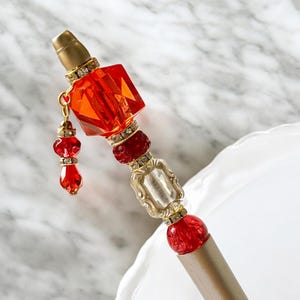 Puede incluir: Un bolígrafo dorado con un adorno de cristal rojo y transparente. El bolígrafo tiene un dije colgante de cristal rojo y transparente.