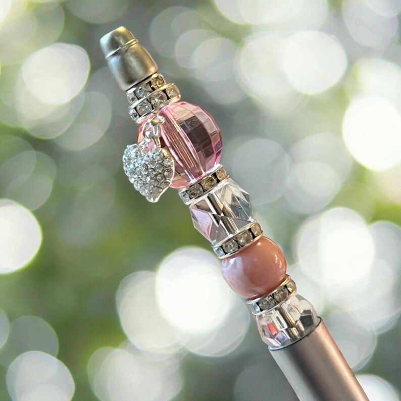 May include: A silver pen with pink and clear beads and rhinestones. A silver heart charm with rhinestones hangs from the pen.