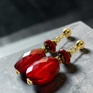 Puede incluir: Un par de pendientes de cristal rojo facetado con detalles dorados. Los pendientes presentan un perno de bola dorada, una pequeña cuenta roja y una cuenta facetada roja más grande y alargada. Los pendientes se muestran sobre una superficie gris.