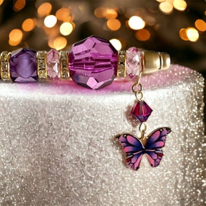 May include: A purple and gold beaded pen with a dangling butterfly charm. The butterfly is purple and pink with gold accents. The pen is on a sparkly silver surface.