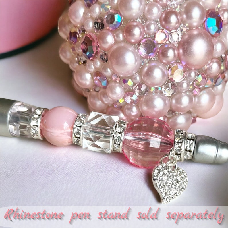 May include: A pink and silver rhinestone pen with a heart-shaped charm. The pen has a clear crystal bead and a pink bead. The pen is lying on a white surface. Rhinestone pen stand sold separately.