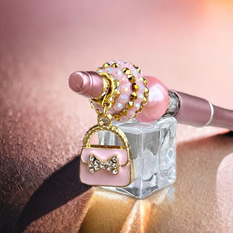 K&ouml;nnte beinhalten: Ein rosa und goldener Anh&auml;nger mit einem kleinen rosa Taschenmotiv h&auml;ngt an einem rosa und goldenen Perlenanh&auml;nger. Der Anh&auml;nger ist an einem rosa und goldenen Stift befestigt.