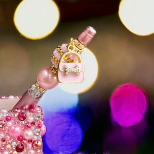 K&ouml;nnte beinhalten: Ein rosa und goldener Stift mit einem Anh&auml;nger in Form einer rosa Handtasche mit einer goldenen Schleife und einem Strassstein. Der Stift ist mit rosa und klaren Strasssteinen verziert.