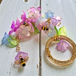 以下が含まれることがあります： ピンク、紫、青の花、緑の葉、金のアクセントが特徴のフローラルキーホルダー。キーホルダーには、透明なラインストーンが付いたゴールドリングと、ぶら下がっている蜂のチャームが含まれています。
