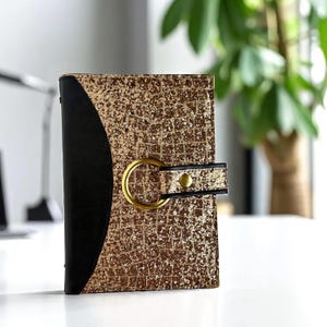 May include: A gold and black journal with a textured, glittery cover. The journal has a gold ring closure and a black spine. The cover has a grid pattern.