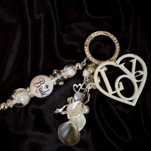 May include: A bridal keychain set with a white heart-shaped charm spelling "LOVE" and a "Bride" bead. The set includes a silver-colored clasp, clear beads, and decorative charms. The background is a black fabric.