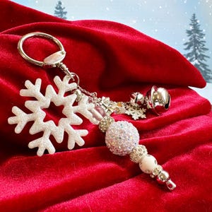 May include: A silver keychain with a white snowflake charm, a cluster of decorative beads, and a small silver bell. The beads include a large, glittery sphere and smaller, sparkling accents. The background is a deep red velvet.