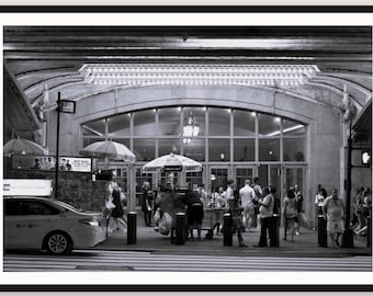 May include: A black and white photograph of a New York City street scene featuring a Sabrett hot dog cart under an umbrella, a parked sedan, and pedestrians walking in front of a large arched building entrance with glass windows and doors.