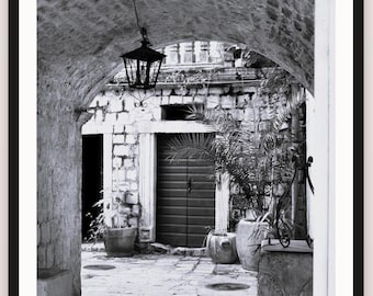 May include: A black and white photograph of a stone archway leading into a courtyard. A hanging lantern is suspended from the ceiling of the arch. The courtyard features a dark wooden door, stone walls, potted plants, and a stone paved floor.
