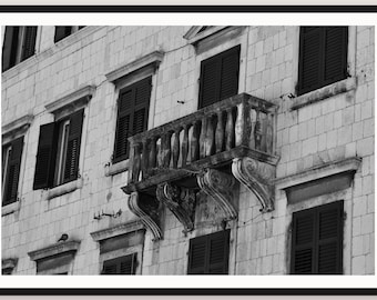 May include: A black and white architectural view of a stone building facade featuring a central balcony with ornate stone balusters and decorative corbels, surrounded by multiple rectangular windows with dark, louvered shutters.