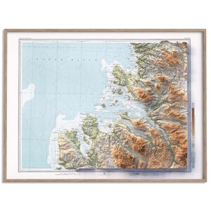 May include: A framed 3D topographic map featuring a light blue ocean area on the left and a detailed, textured landmass on the right with brown mountain ranges and green coastal regions, all enclosed within a simple light wood frame.