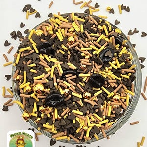 May include: A clear glass bowl filled with a mix of decorative sprinkles. The sprinkles include dark brown heart shapes, yellow sticks, tan sticks, gold spheres, and black coffee bean shapes. The bowl is surrounded by loose sprinkles.