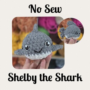 May include: A gray and white crocheted shark toy with large, round eyes and a smiling mouth. The text "No Sew" and "Shelby the Shark" are displayed above and below the shark. A smaller version of the shark is shown in the upper right corner.