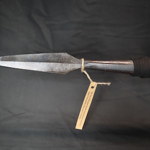 May include: A long, silver spear with a black leather wrapped handle. The spear has a sharp point and a tag attached to the handle that reads "The tip of the spear, I went to war, and I came home."