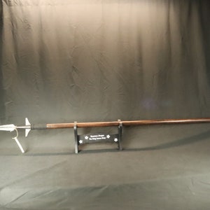 May include: A silver spear with a wooden shaft, mounted on a black stand. The stand has the text "Custom Blades by The Holy Grail Shop" printed on it.