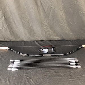 May include: A black recurve bow with white tips, a black arrow rest, and a set of arrows with white fletching. The bow is lying on a dark surface.