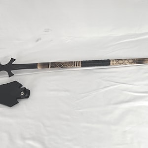 May include: A black spear with a silver blade and decorative handle. The handle has a black grip with gold-colored accents and a sheath. The spear is lying on a white surface.