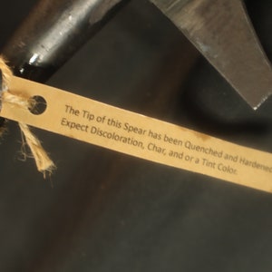 Peut inclure: Gros plan d'une pointe de lance en m&eacute;tal sombre avec une &eacute;tiquette attach&eacute;e. La pointe est tranchante et angulaire. L'&eacute;tiquette indique : "The Tip of this Spear has been Quenched and Hardened. Expect Discoloration, Char, and or a Tint Color."