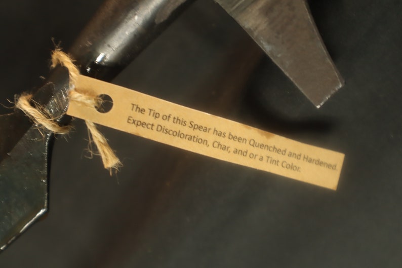 Peut inclure: Gros plan d'une pointe de lance en m&eacute;tal sombre avec une &eacute;tiquette attach&eacute;e. La pointe est tranchante et angulaire. L'&eacute;tiquette indique : "The Tip of this Spear has been Quenched and Hardened. Expect Discoloration, Char, and or a Tint Color."