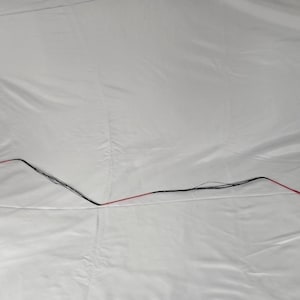 May include: A black and red whip, lying on a white surface. The whip is thin and flexible, with a red handle and a black body. The whip is designed for use in equestrian activities.