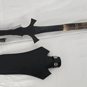 May include: A black spear with a decorative handle and a matching black sheath. The spearhead is silver and black with a unique, jagged design. The handle features a carved pattern and leather wrapping.