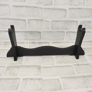 May include: A black wooden sword stand with two slots for displaying swords. The stand has a curved base and is made of solid wood.