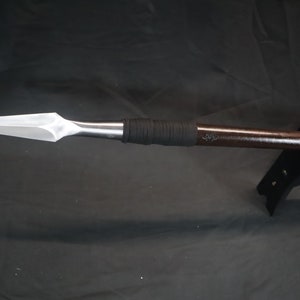 May include: A silver spearhead with a dark brown wooden shaft and black leather grip. The spear is mounted on a black stand with a white logo that reads "Guardian Dagger The Shop That Makes Things Sharp".