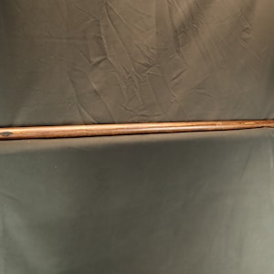 Peut inclure: Un long b&acirc;ton en bois avec une finition sombre. Le b&acirc;ton a une poign&eacute;e envelopp&eacute;e pr&egrave;s du haut et se r&eacute;tr&eacute;cit en pointe. Le grain du bois est visible, et le b&acirc;ton semble fait &agrave; la main. Le fond est un tissu sombre.