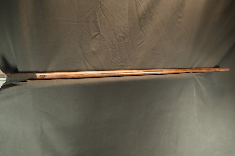 Peut inclure: Un long b&acirc;ton en bois avec une finition sombre. Le b&acirc;ton a une poign&eacute;e envelopp&eacute;e pr&egrave;s du haut et se r&eacute;tr&eacute;cit en pointe. Le grain du bois est visible, et le b&acirc;ton semble fait &agrave; la main. Le fond est un tissu sombre.