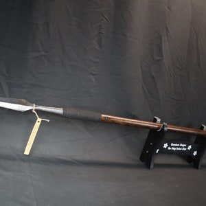 May include: A long, wooden-handled spear with a metal tip and a black leather wrap. The spear is mounted on a black stand with the text "Guardian Shoppe The Only Samurai Shop" printed on it.